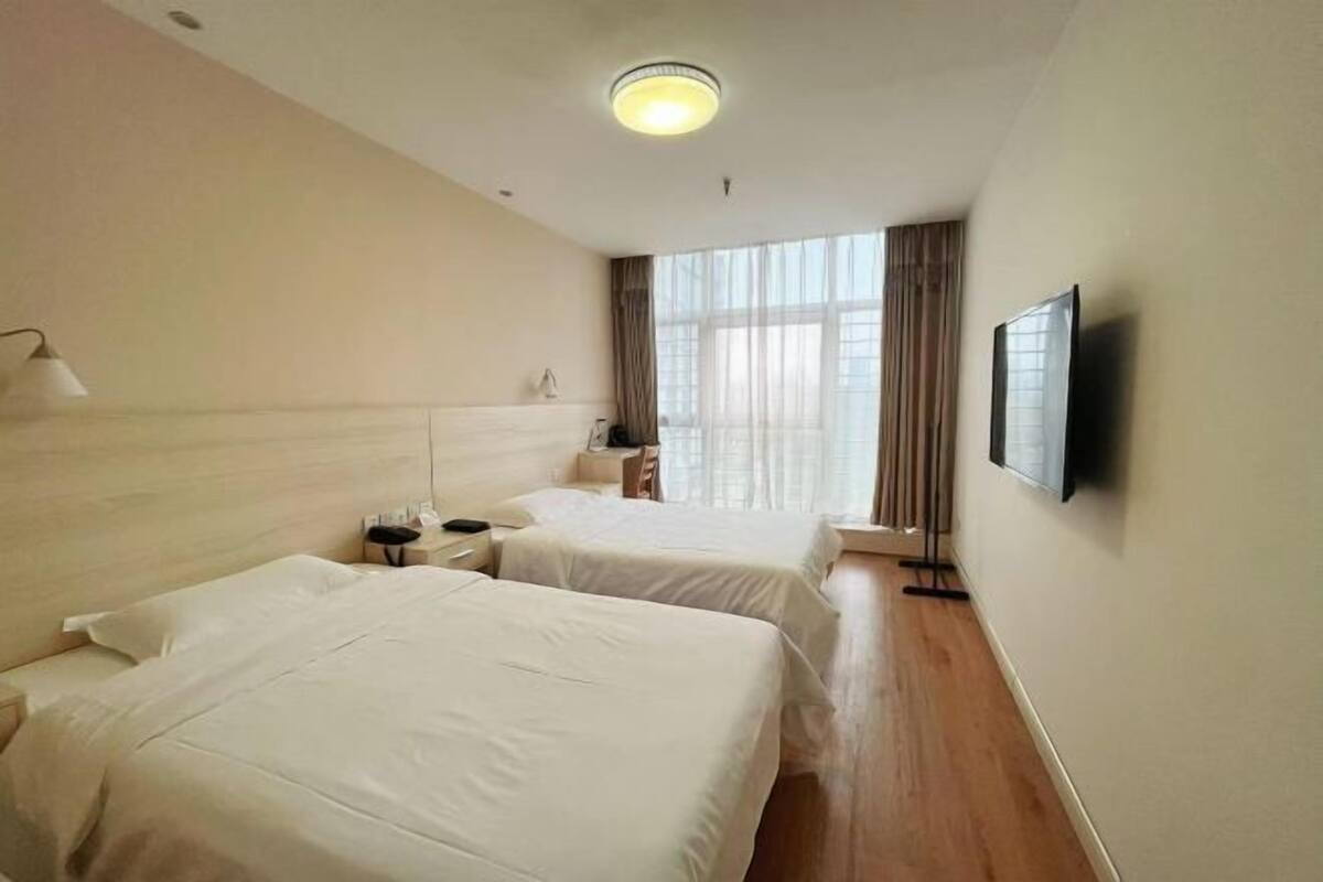 Ein Hotelzimmer mit zwei Betten, einem Flachbildfernseher, einem Nachttisch mit Lampe und einem Fenster mit Vorhängen.