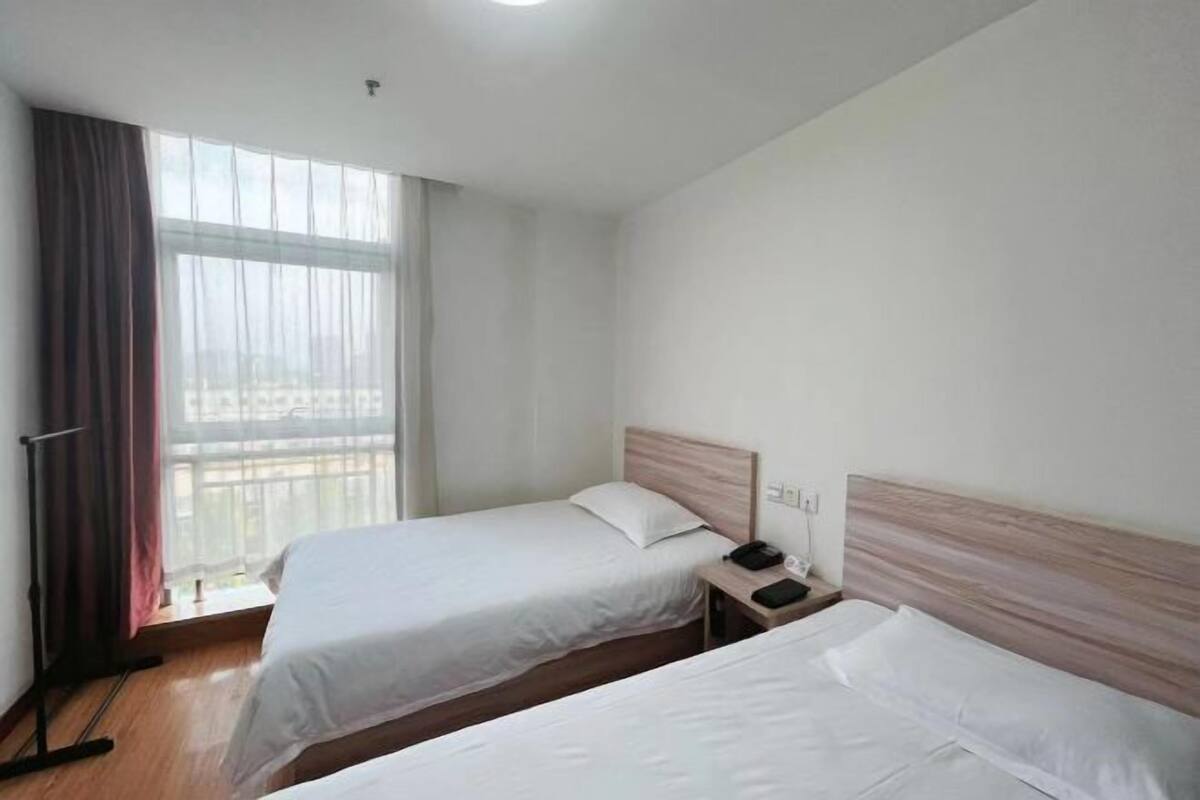 Ein Hotelzimmer mit zwei Einzelbetten, einem Fenster mit Vorhängen, Holzboden und einem Nachttisch mit Telefon und Lampe.