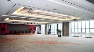 A spacious conference room with a carpeted floor, a stage with a screen, and large windows providing natural light.