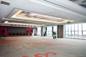 A spacious conference room with a carpeted floor, a stage with a screen, and large windows providing natural light. - Guanhe International Hotel (Shijiazhuang)