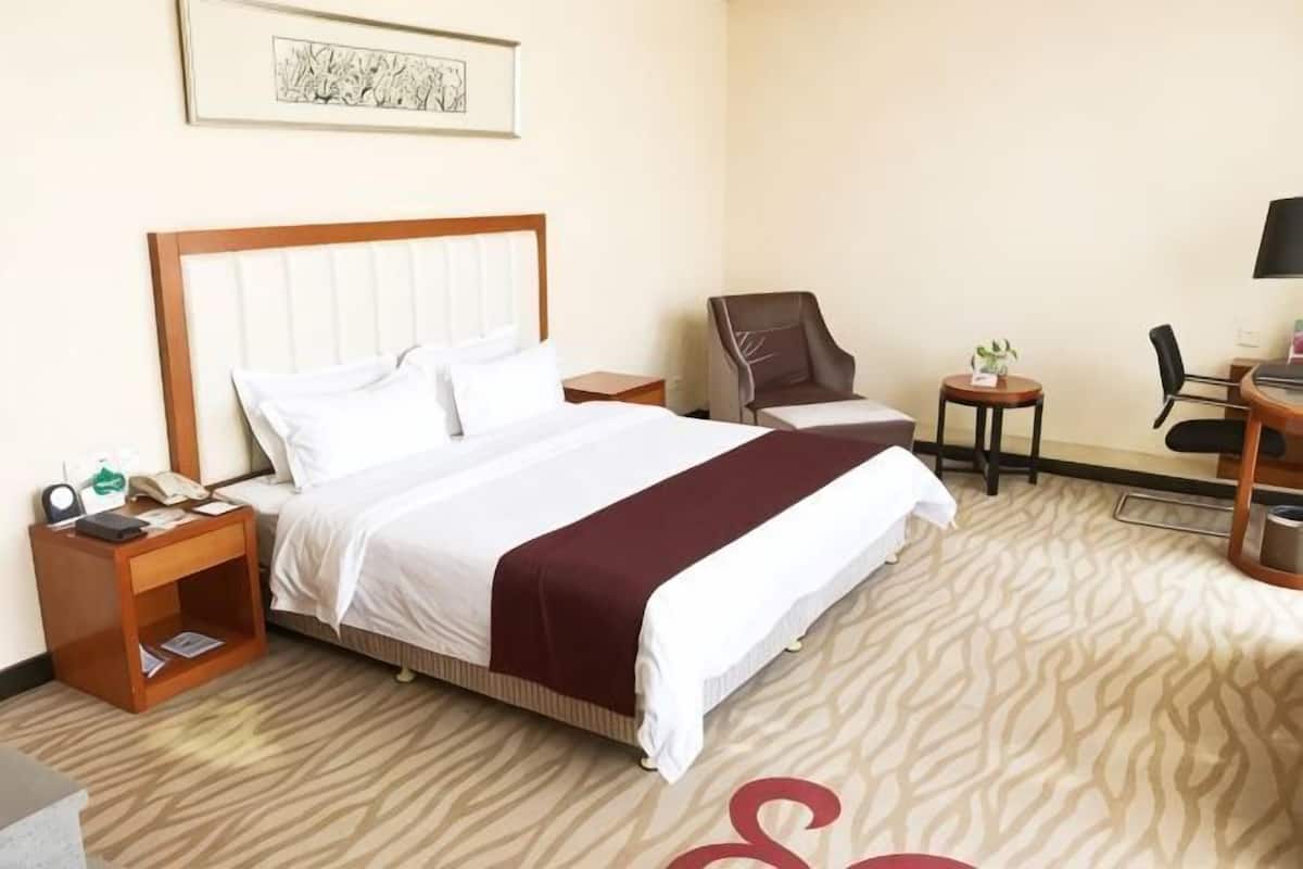 Ein Hotelzimmer mit einem großen Bett, einem Schreibtisch, einem Sessel, einem kleinen Tisch mit Pflanze und einem gerahmten Bild an der Wand.