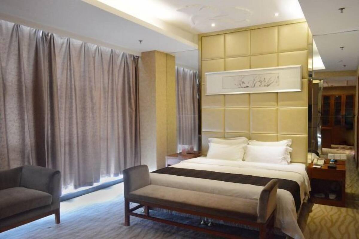 Ein Hotelzimmer mit Bett, Sessel, Nachttisch und einem großen Fenster mit Vorhängen.