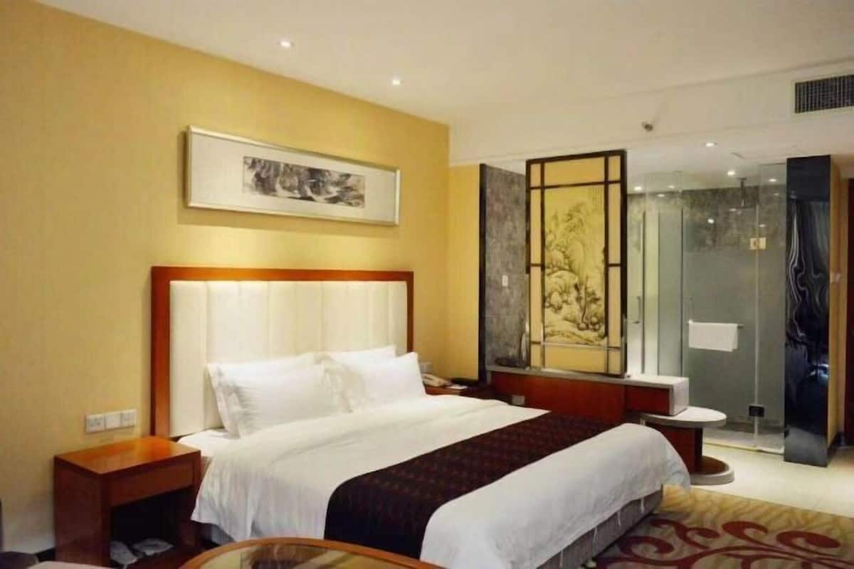 Ein Hotelzimmer mit einem großen Bett, einem Nachttisch, einer Duschkabine aus Glas und einem gerahmten Kunstwerk an der Wand.