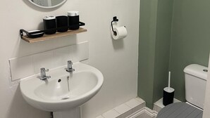 A bathroom with a white sink, a mirror, a toilet, and a shelf with toiletries. - Family Friendly One Bedroom Apartment w King Bed (Colchester)