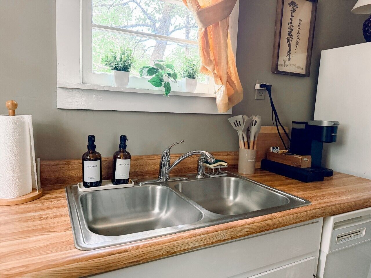 A kitchen with a double sink, wooden countertop, and a window with plants.