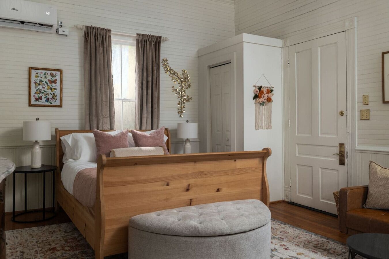 A bedroom with a wooden bed, bedside lamps, a grey ottoman, a floral macrame wall hanging, and a door with a brass handle.