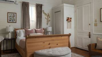 A bedroom with a wooden bed, bedside lamps, a grey ottoman, a floral macrame wall hanging, and a door with a brass handle.