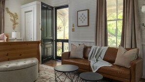 Una sala de estar acogedora con un sofá de cuero marrón, un reposapiés gris y una mesa de centro redonda. Un banco de madera está apoyado contra la pared, y una puerta da a un espacio al aire libre.