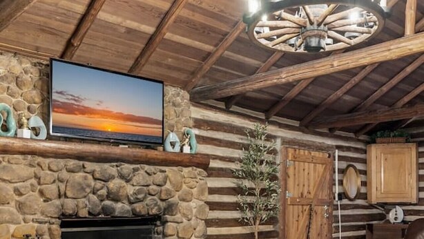A rustic cabin interior with a stone fireplace, leather sofa, wooden beams, and a mounted TV. - The Cabin - Fire Pit, Pool Table, Spacious (Greensboro)