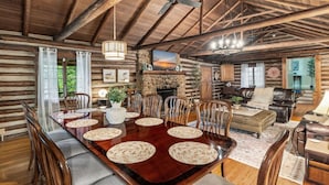 A rustic cabin interior with a wooden dining table surrounded by chairs, a stone fireplace, and a cozy seating area with leather sofas. - The Cabin - Fire Pit, Pool Table, Spacious (Greensboro)