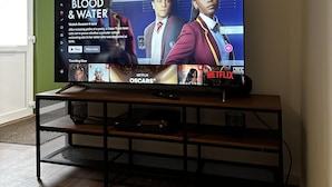 A flat-screen TV on a wooden stand in a room with a green wall and wooden flooring. - Entire Apartment Super King Bed Close To Town (Colchester)