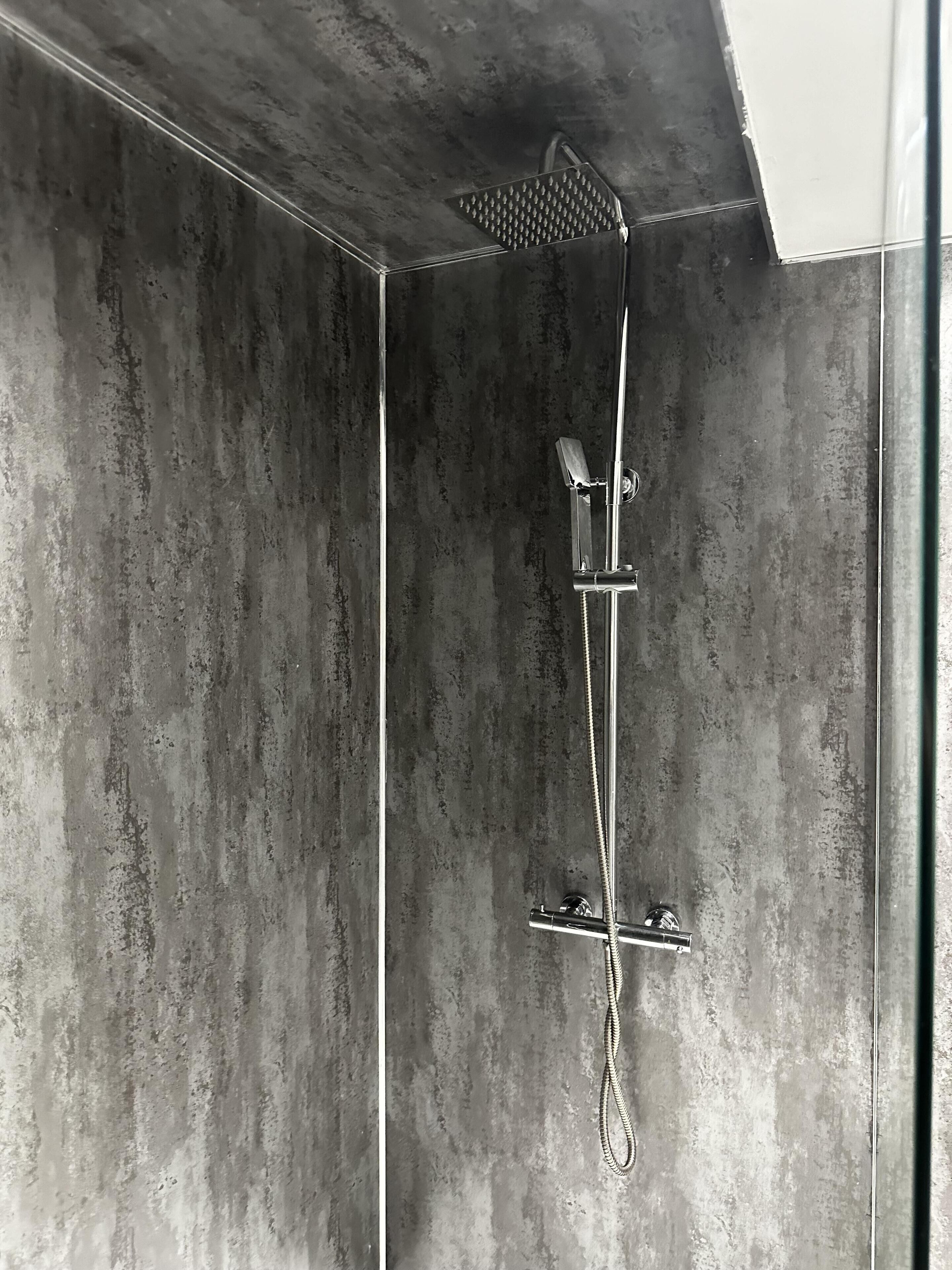 Une douche moderne dotée d’un pommeau de douche à main et d’une grande pomme de douche au plafond, le tout installé sur un mur à texture.