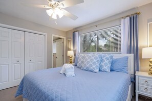 A bedroom with a bed, a ceiling fan, a window with blinds, and a nightstand with a lamp.