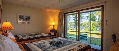 A hotel room with two beds, a chair, a table, a lamp, and a large sliding glass door leading to a balcony with a view of a lake.