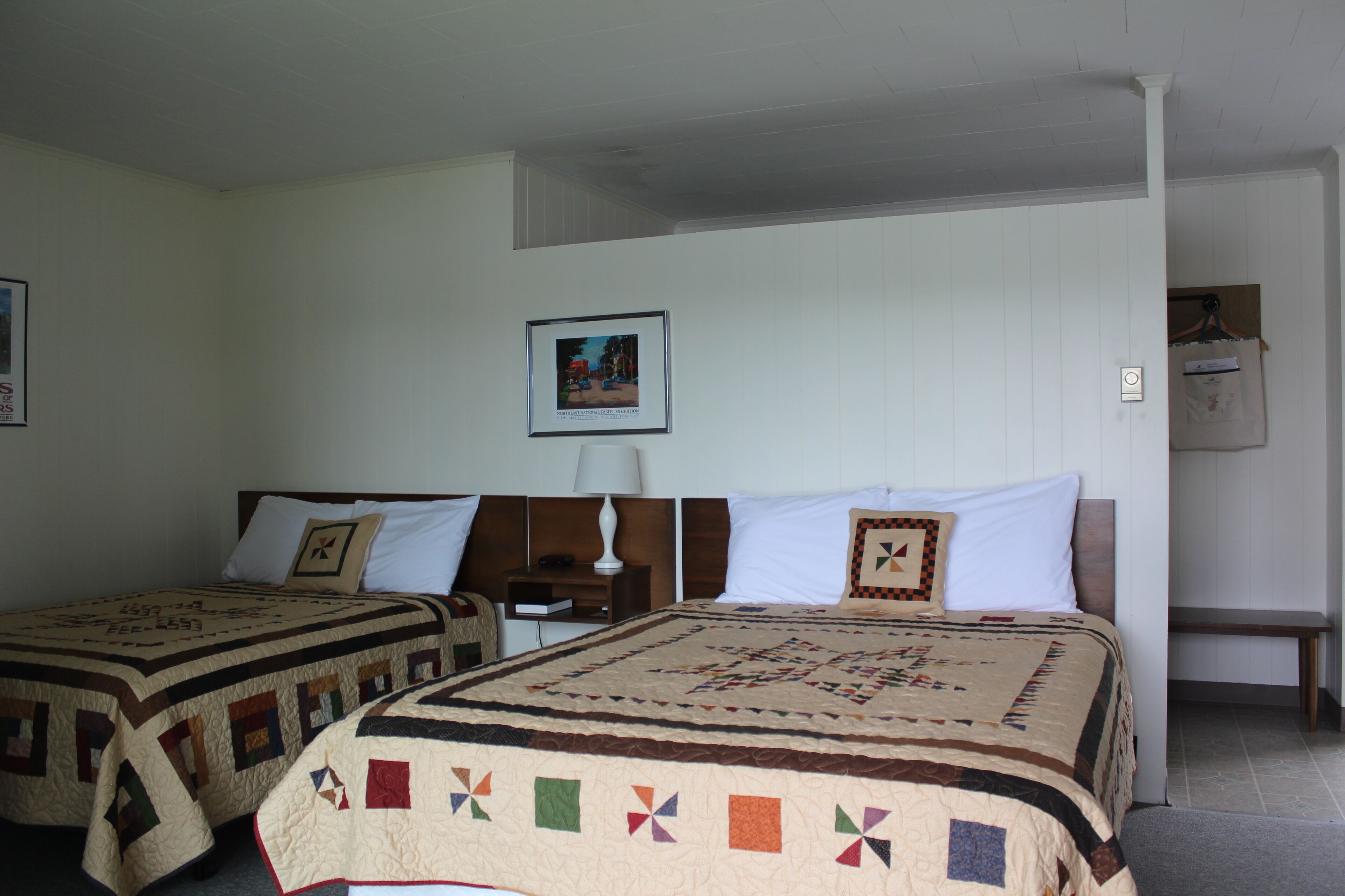 Una stanza con due letti, ognuno con una coperta, un comodino con una lampada, un quadro appeso al muro e una panca nell'angolo.