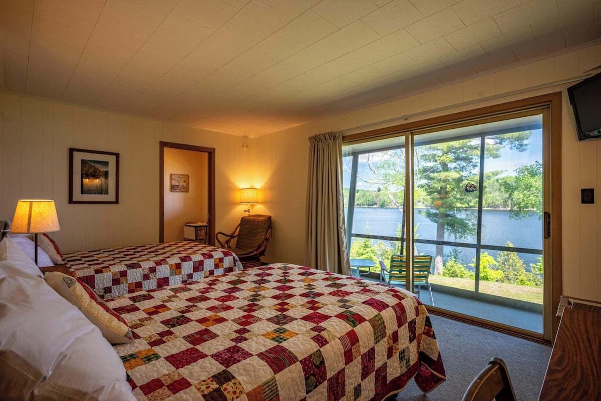 A hotel room with two beds, a chair, a table, and a large window offering a view of a lake.