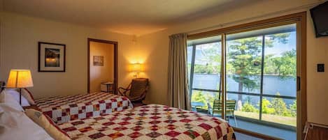 A hotel room with two beds, a chair, a table, and a large window offering a view of a lake.