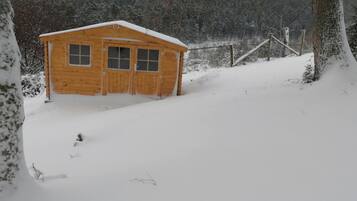 En stuga av trä med två dörrar och fönster, omgiven av snötäckta träd och ett staket.