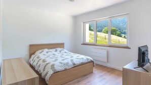A bedroom with a bed, a desk, a radiator, and a window with a view of a green landscape.