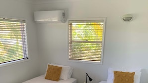 Ein Hotelzimmer mit zwei Einzelbetten, jeweils mit grauer Bettdecke, weißen Kissen und einem aufgerollten weißen Handtuch. Es gibt zwei Fenster mit Jalousien, über jedem Fenster eine Klimaanlage sowie einen kleinen Tisch mit Lampe und einer Topfpflanze zwischen den Betten.