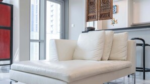 Un sofá de cuero blanco con estructura metálica, ubicado en una habitación moderna con ventanas amplias, una pared de acento roja y paneles decorativos de madera.