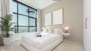 A modern bedroom with a large bed, a potted plant, and a view through a large window. - StayShort-Elegant 1 Bedroom City View Apartment Square Tower JVC (Dubai)