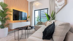 A modern living room with a large flat-screen TV, a beige sofa, a coffee table, and a potted plant. - StayShort-Elegant 1 Bedroom City View Apartment Square Tower JVC (Dubai)