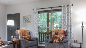 Un salon avec deux fauteuils gris, une table basse en bois, un lampadaire et une fenêtre ornée de rideaux à motifs.