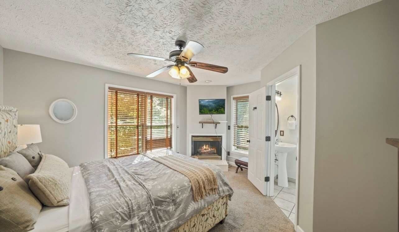 A bedroom with a bed, a ceiling fan, a fireplace, and a view of the outdoors.