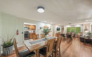 Een ruime woonkamer met een eettafel, stoelen en een bank. Er is een keuken met kastjes en een fornuis.