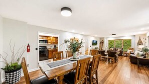 A spacious living area with a wooden dining table, comfortable seating, a kitchen in the background, and a large window with a view.