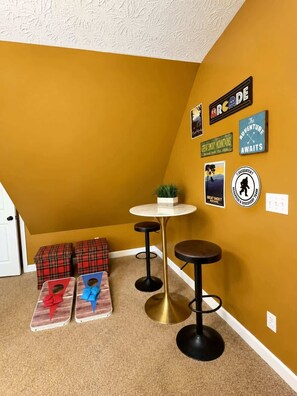 Ein Zimmer mit gelber Wand, weißer Decke, einem runden Tisch mit goldener Basis, zwei schwarzen Hocker, karierten Aufbewahrungsboxen und Wanddekorationen, darunter ein Schild mit der Aufschrift 'The Adventure Awaits'.