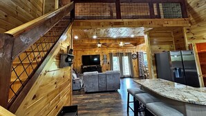 Un intérieur de cabine spacieux avec un escalier en bois, un coin salon doté d’un canapé et d’une télévision, une cuisine avec un plan de travail en marbre et un coin repas avec des tabourets de bar.