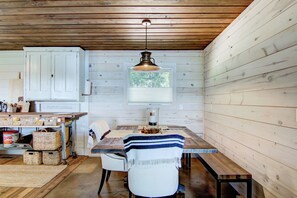 Interior rústico de una cabaña con una mesa de madera, sillas blancas, una lámpara colgante y una ventana con vistas a los árboles.
