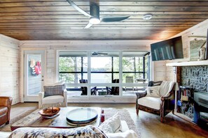 A cozy cabin interior with wooden walls, a ceiling fan, a large window overlooking a lake, and a fireplace. - Lakefront Living at its Finest at Clearwater Cove on LCO (Hayward)