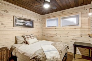 Un dormitorio con paneles de madera, una cama, almohadas, una colcha, un cuadro enmarcado y dos ventanas.