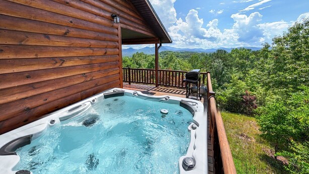 A wooden cabin with a hot tub on a deck overlooking a forested landscape. - Sunsets and Moonshine Log Cabin with Outstanding Mountain Views (Bryson City)