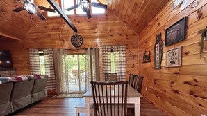 A wooden cabin interior with a dining table, chairs, a sofa, a ceiling fan, and a clock.