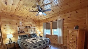 A wooden cabin bedroom with a bed, a nightstand, a dresser, a ceiling fan, and two windows with curtains.