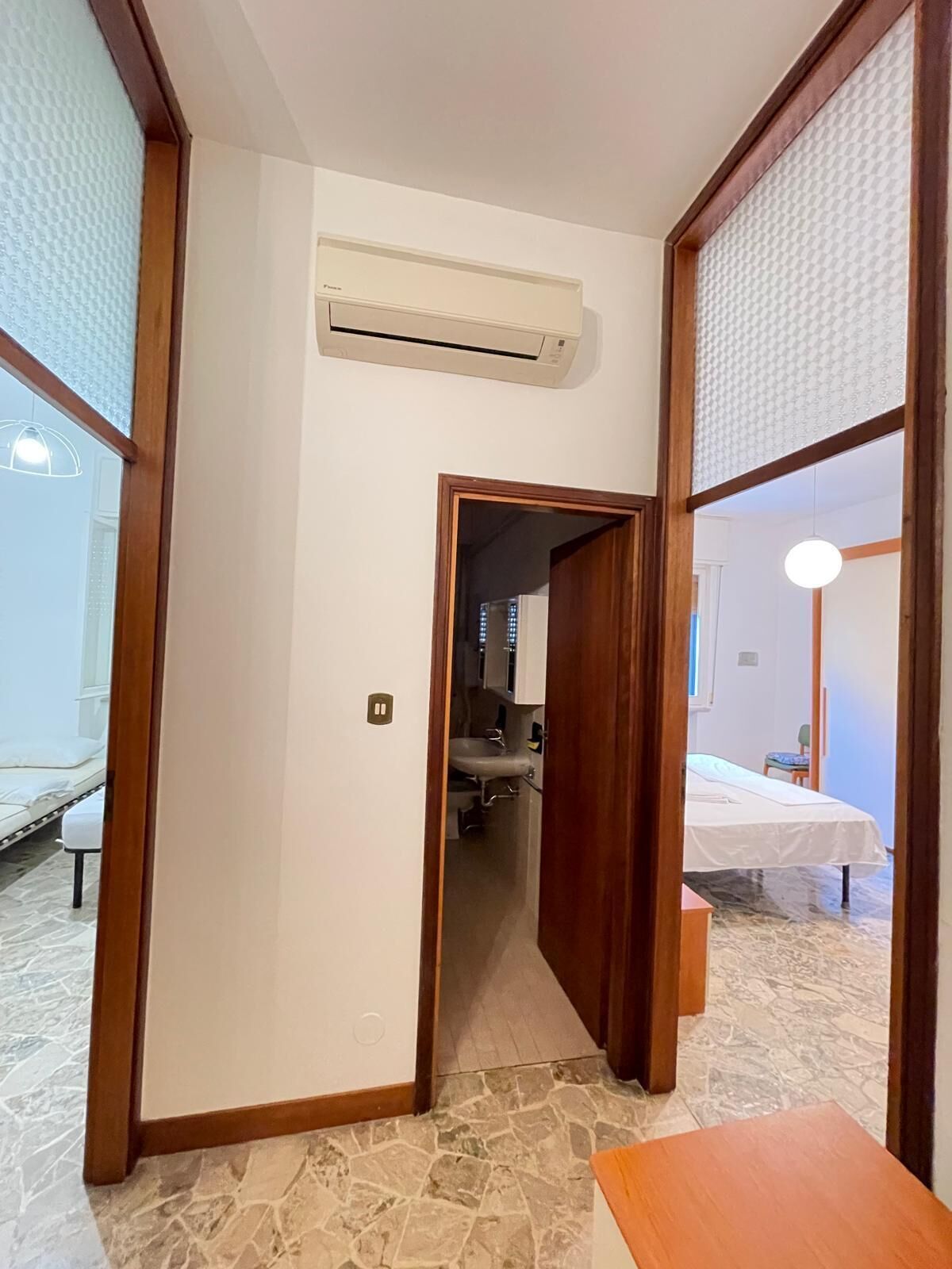 A hotel room with a stone-tiled floor, a bed, a bedside table, a wooden door, and an air conditioning unit.
