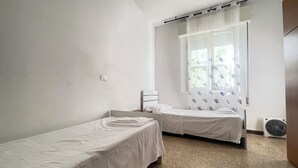 A room with two single beds, a window with heart-patterned curtains, a standing fan, and a light switch on the wall.