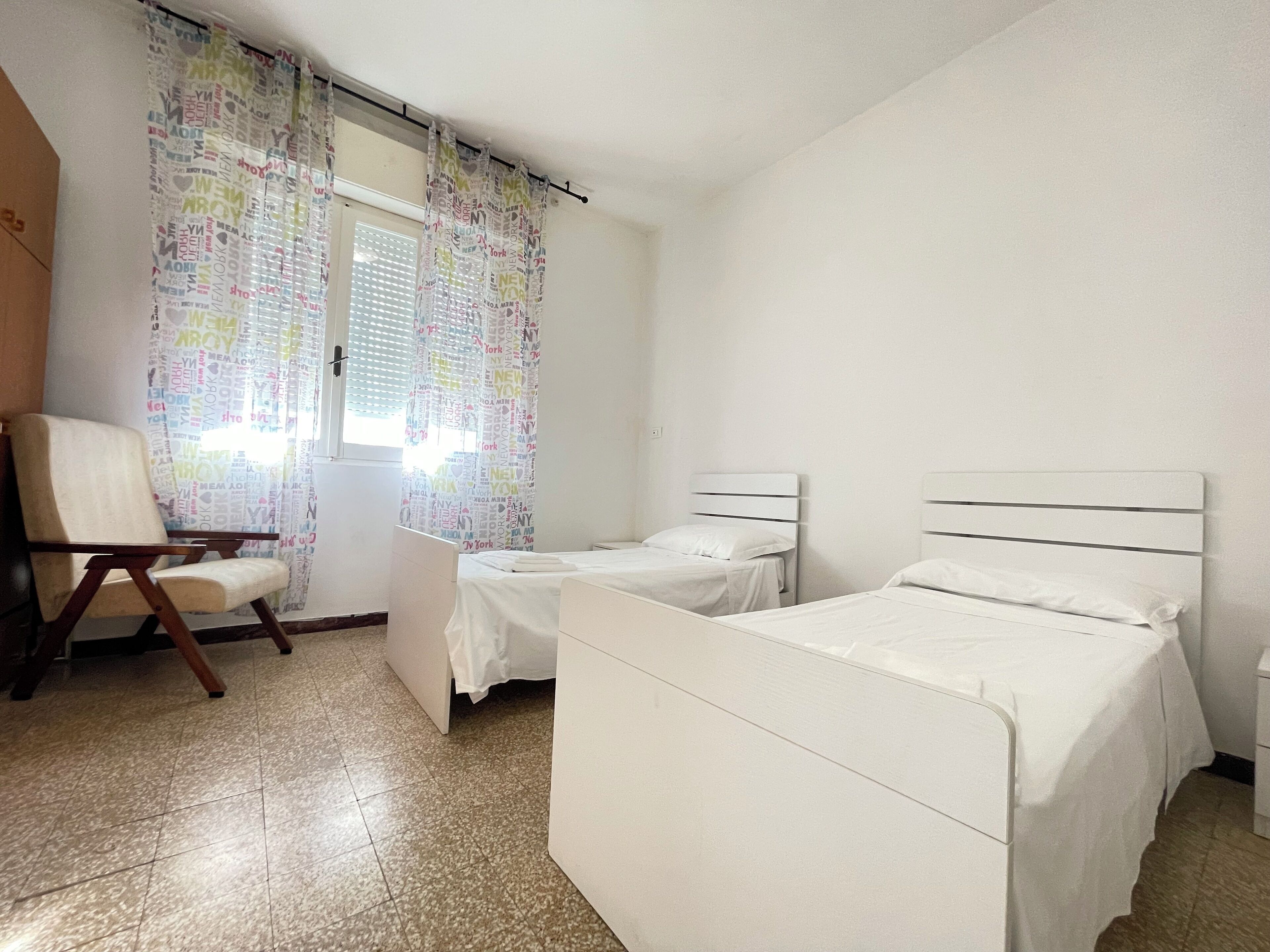 A room with two single beds, a chair, and a window with patterned curtains.