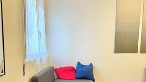A small, modern room with a grey sofa, a red and a blue pillow, a window with sheer curtains, and a large abstract painting on the wall.