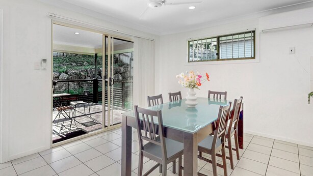 A dining area with a glass table, wooden chairs, a ceiling fan, and a window with blinds.