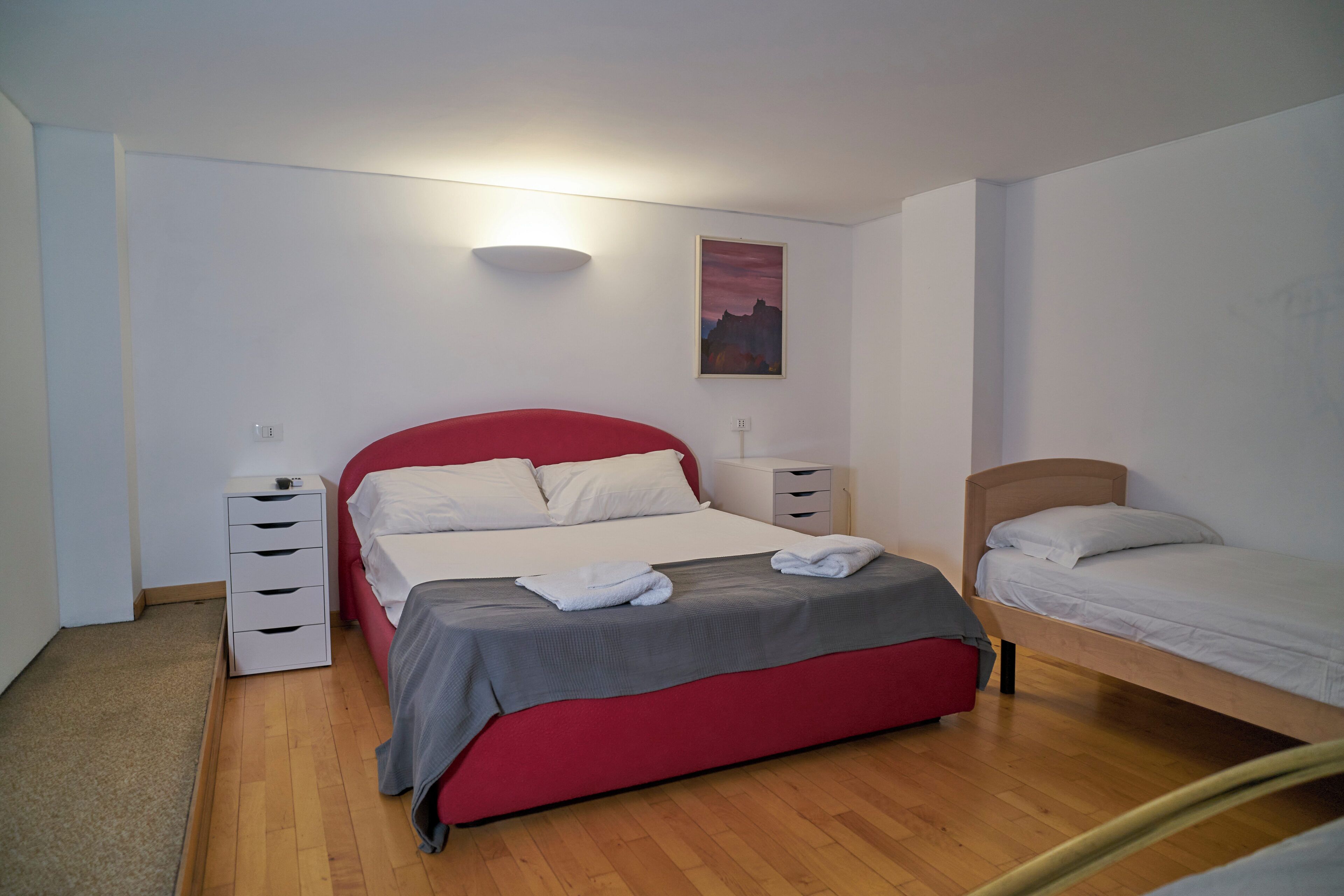 Een slaapkamer met een rood bed, een kleiner bed, een nachtkastje, een ladekast en een wandlamp.