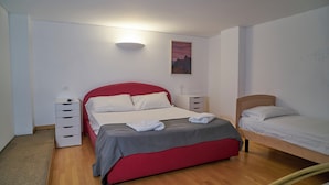 Une chambre avec un lit rouge, un lit plus petit, une table de chevet, une commode et une lampe fixée au mur.