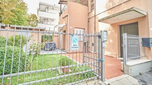 Edificio residencial con acceso restringido, un pequeño jardín, una puerta visible y un letrero que indica zona de no estacionamiento.