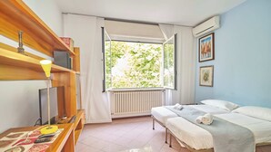 Habitación de hotel con una cama, un escritorio con lámpara, un radiador y una ventana con cortinas.