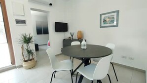 Een eetruimte met een ronde tafel, witte stoelen, een potplant, een decoratieve vaas en een lijst met een afbeelding aan de muur.