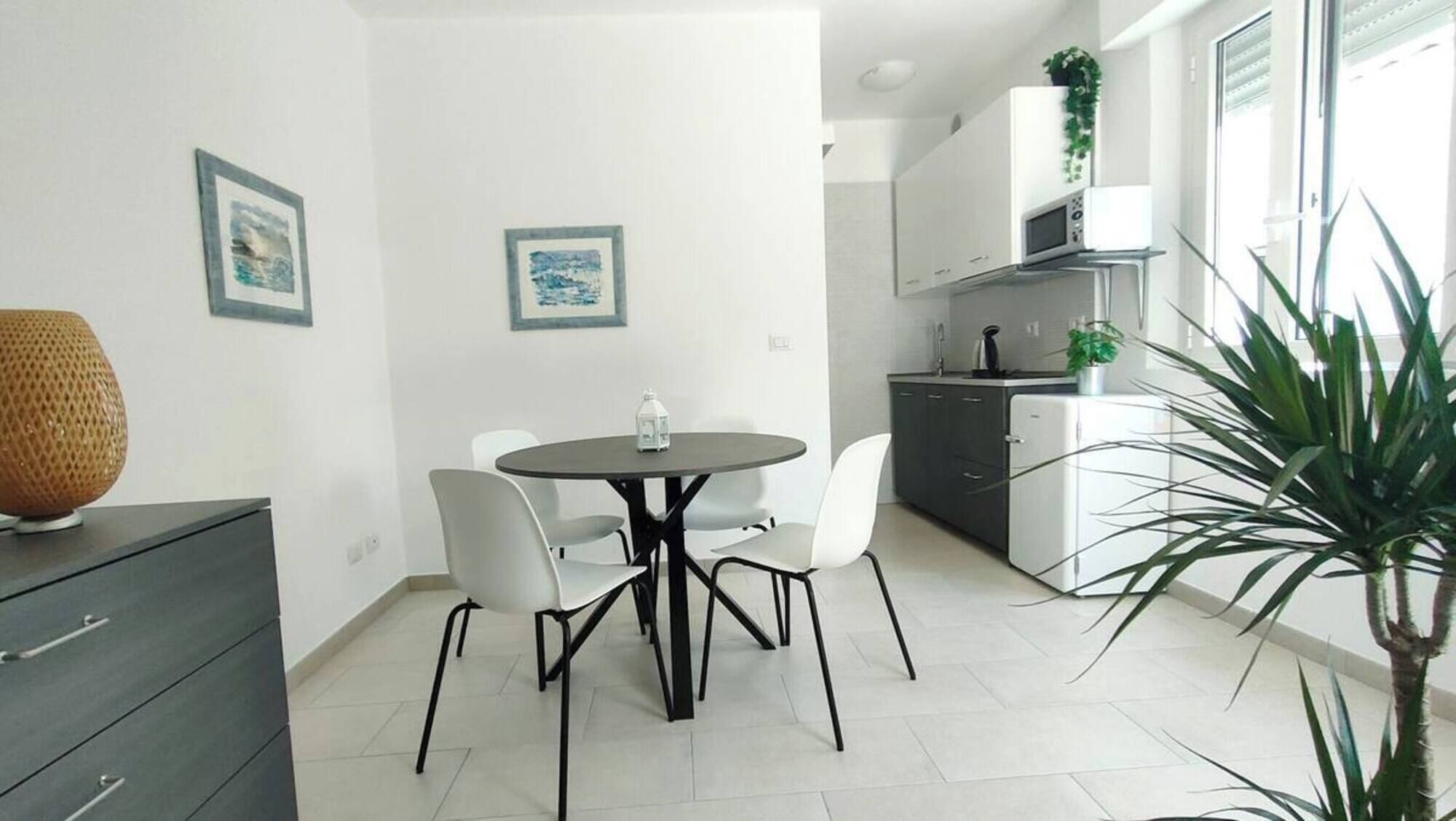 A modern kitchen with white cabinets, a microwave, and a small refrigerator. There is a dining table with chairs and a potted plant.